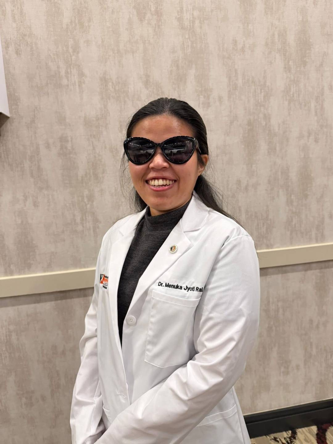 Wearing sunglasses and a white coat with her name embroidered on it, Menuka smiles.